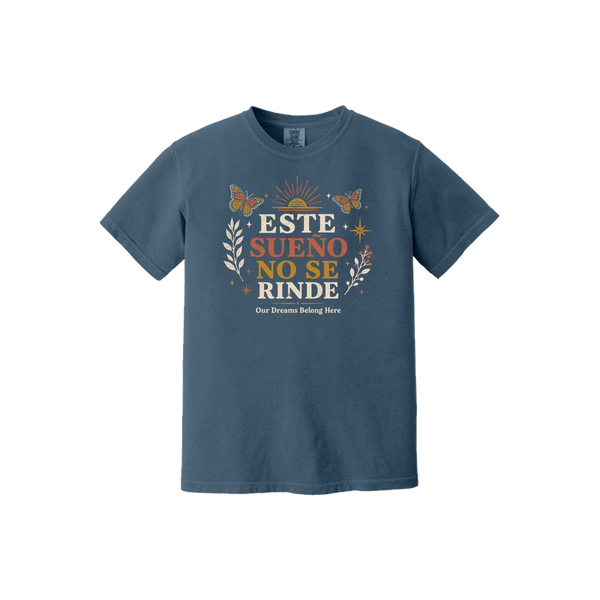 A slate blue short-sleeve t-shirt with a colorful graphic on the chest that reads 'ESTE SUENO NO SE RINDE' with butterflies, sun rays, and leaves, displayed on a solid white background.