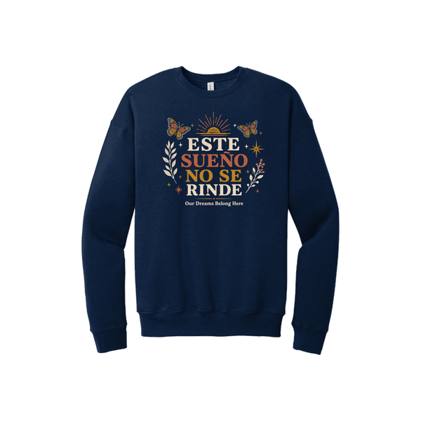 A navy blue crewneck sweatshirt with a colorful graphic on the chest featuring the Spanish text 'ESTE SUENO NO SE RINDE' in orange and white, surrounded by butterflies, stars, and floral designs, with the smaller text 'Our Dreams Belong Here' underneath.