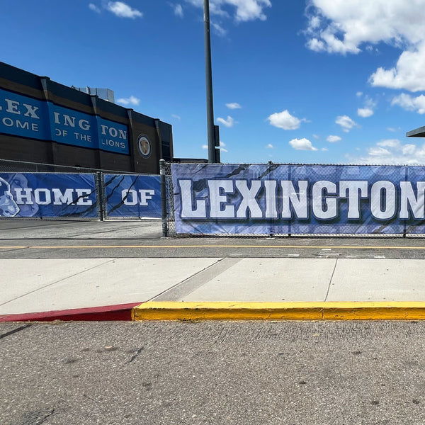 A blue privacy mesh banner reading 'HOME OF LEXINGTON' is displayed on a chain-link fence in an outdoor setting with a sidewalk, asphalt road, and a blue sky with clouds in the background.
